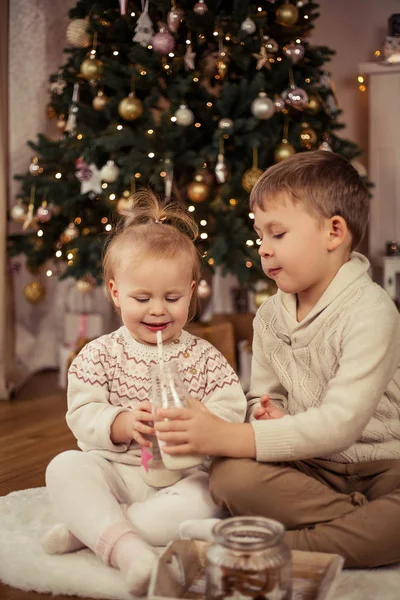 Piccolo ragazzo carino felice e ragazza sedersi vicino all'albero di Natale e bere latte con biscotti di Natale. Natale. Anno nuovo . — Foto Stock