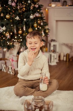 Küçük şirin çocuk bir Noel ağacının yanında oturur ve Noel kurabiyeleriyle süt içer. Noel zamanı. Yeni Yıl.
