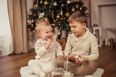 Küçük tatlı mutlu çocuk ve kız Noel ağacının yanında oturur ve Noel kurabiyeleriyle süt içerler. Noel zamanı. Yeni Yıl.