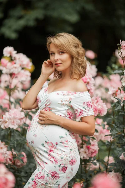 En vacker ung gravid kvinna går i en rosenträdgård. Porträtt av en gravid kvinna i klänning. Sommar. — Stockfoto