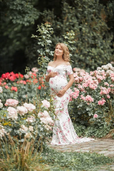 En vacker ung gravid kvinna går i en rosenträdgård. Porträtt av en gravid kvinna i klänning. Sommar. — Stockfoto