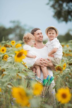 Mutlu bir aile ayçiçeği tarlasında yürüyor. Ayçiçeği tarlasında iki çocuğu olan yakışıklı bir baba..