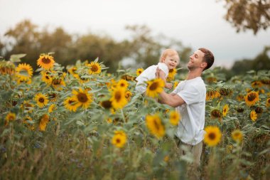 Genç güzel bir kız çocuğunu bir ayçiçeği tarlasında kollarında tutuyor. Mutlu bir aile ayçiçeği tarlasında yürüyor.