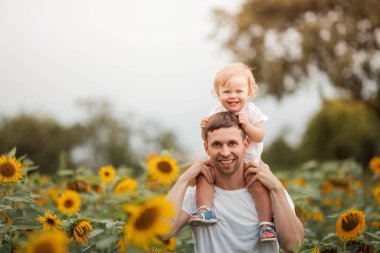 Genç güzel bir kız çocuğunu bir ayçiçeği tarlasında kollarında tutuyor. Mutlu bir aile ayçiçeği tarlasında yürüyor.