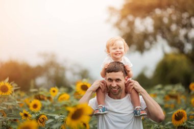Genç güzel bir kız çocuğunu bir ayçiçeği tarlasında kollarında tutuyor. Mutlu bir aile ayçiçeği tarlasında yürüyor.