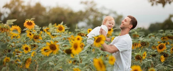 若い美しいひまわり畑で彼女の腕の中で小さな娘を保持しています。幸せな家族がひまわり畑を歩いています. — ストック写真
