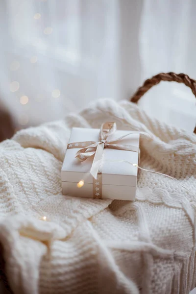 Piccola confezione regalo bianca con nastro su un maglione lavorato a maglia, cesto di vimini, luci di Natale . — Foto Stock