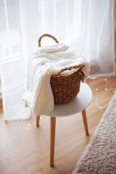 Uma cesta de vime com linho fica na janela. Camisola de malha, luzes de Natal. Aconchegante . — Fotografia de Stock