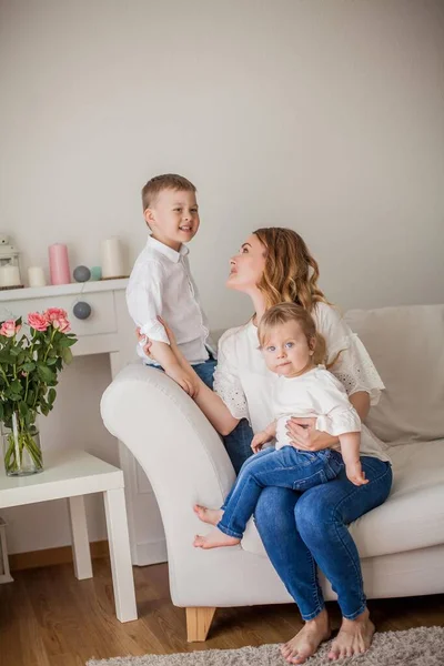 Gyönyörű fiatal anya, kisfiú, kislánya ül a kanapén otthon. Anyák napja. Egy boldog család. Hangulatos. — Stock Fotó