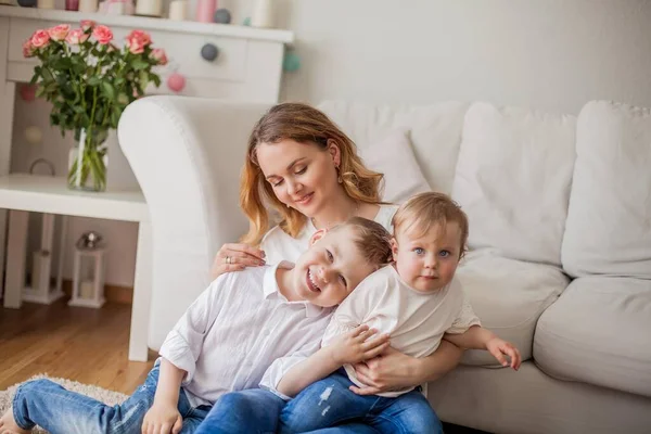 Mooie jonge moeder, kleine zoon, kleine dochter zitten op de Bank thuis. Moederdag. Een gelukkige familie. Gezellige. — Stockfoto