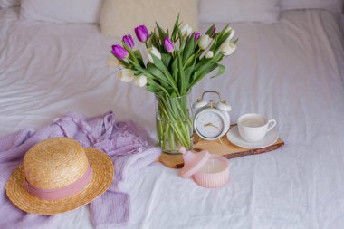 A bouquet of tulips in a glass vase, a white alarm clock, a candle, a note, a cappuccino mug on a wooden board are standing on the bed. Womens things. Breakfast in bed.