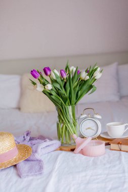 A bouquet of tulips in a glass vase, a white alarm clock, a candle, a note, a cappuccino mug on a wooden board are standing on the bed. Womens things. Breakfast in bed.