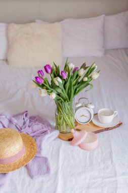 A bouquet of tulips in a glass vase, a white alarm clock, a candle, a note, a cappuccino mug on a wooden board are standing on the bed. Womens things. Breakfast in bed.