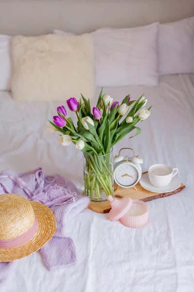 A bouquet of tulips in a glass vase, a white alarm clock, a candle, a note, a cappuccino mug on a wooden board are standing on the bed. Womens things. Breakfast in bed.