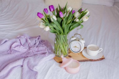 A bouquet of tulips in a glass vase, a white alarm clock, a candle, a note, a cappuccino mug on a wooden board are standing on the bed. Womens lace underwear. Breakfast in bed.