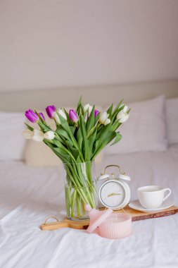 A bouquet of tulips in a glass vase, a white alarm clock, a candle, a note, a cappuccino mug on a wooden board are standing on the bed. Spring. Breakfast in bed.