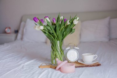 A bouquet of tulips in a glass vase, a white alarm clock, a candle, a note, a cappuccino mug on a wooden board are standing on the bed. Spring. Breakfast in bed.