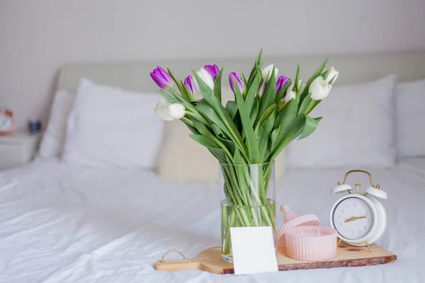 Un mazzo di tulipani in un vaso di vetro, una sveglia bianca, una candela, una nota su una tavola di legno poggiano sul letto. Primavera . — Foto Stock