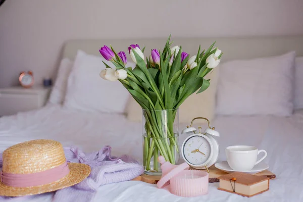 Un mazzo di tulipani in un vaso di vetro, una sveglia bianca, una candela, una nota, una tazza di cappuccino su una tavola di legno sono in piedi sul letto. Cose da donne. Colazione a letto . — Foto Stock