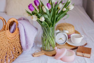 A bouquet of tulips in a glass vase, a white alarm clock, a candle, a note, a cappuccino mug on a wooden board are standing on the bed. Womens things. Breakfast in bed.