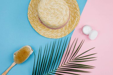 Tropical plant, womens straw hat, cream in a jar, body brush made of natural materials on a blue with pink background.