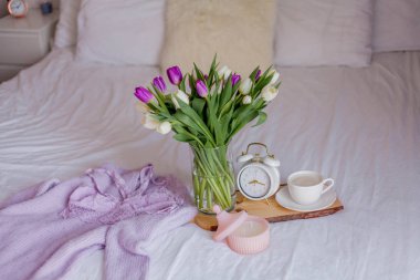 A bouquet of tulips in a glass vase, a white alarm clock, a candle, a note, a cappuccino mug on a wooden board are standing on the bed. Womens lace underwear. Breakfast in bed.