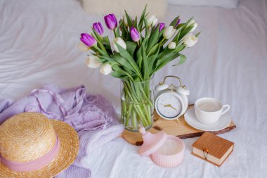 A bouquet of tulips in a glass vase, a white alarm clock, a candle, a note, a cappuccino mug on a wooden board are standing on the bed. Womens things. Breakfast in bed.