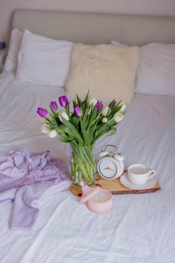A bouquet of tulips in a glass vase, a white alarm clock, a candle, a note, a cappuccino mug on a wooden board are standing on the bed. Women's lace underwear. Breakfast in bed.