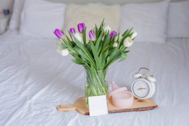 A bouquet of tulips in a glass vase, a white alarm clock, a candle, a note on a wooden board stand on the bed. Spring.