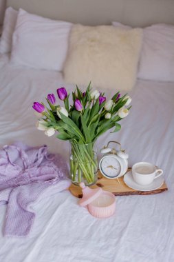 A bouquet of tulips in a glass vase, a white alarm clock, a candle, a note, a cappuccino mug on a wooden board are standing on the bed. Women's lace underwear. Breakfast in bed.
