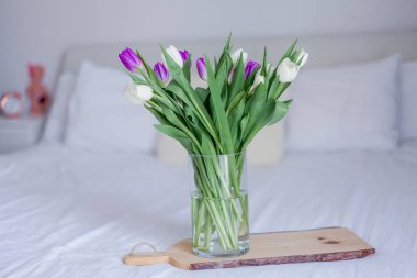 Bouquet of tulips in a glass vase, on a wooden board stand on the bed. Spring.