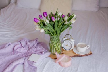 A bouquet of tulips in a glass vase, a white alarm clock, a candle, a note, a cappuccino mug on a wooden board are standing on the bed. Women's lace underwear. Breakfast in bed.
