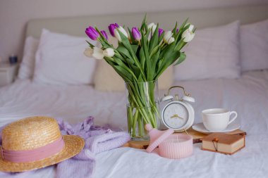 A bouquet of tulips in a glass vase, a white alarm clock, a candle, a note, a cappuccino mug on a wooden board are standing on the bed. Womens things. Breakfast in bed.