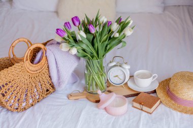 A bouquet of tulips in a glass vase, a white alarm clock, a candle, a note, a cappuccino mug on a wooden board are standing on the bed. Women's things. Breakfast in bed.