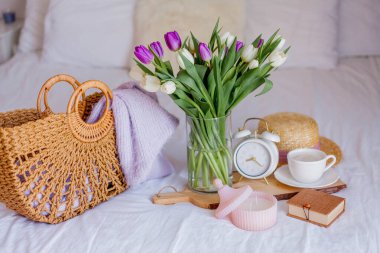 A bouquet of tulips in a glass vase, a white alarm clock, a candle, a note, a cappuccino mug on a wooden board are standing on the bed. Women's things. Breakfast in bed.
