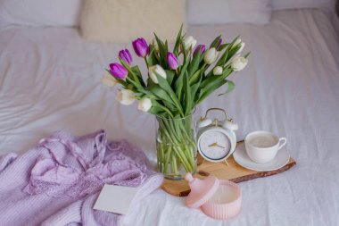 A bouquet of tulips in a glass vase, a white alarm clock, a candle, a note, a cappuccino mug on a wooden board are standing on the bed. Women's lace underwear. Breakfast in bed.
