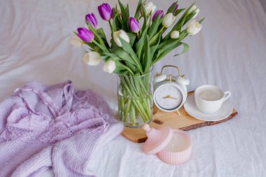 A bouquet of tulips in a glass vase, a white alarm clock, a candle, a note, a cappuccino mug on a wooden board are standing on the bed. Women's lace underwear. Breakfast in bed.