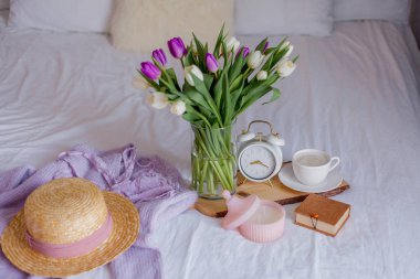 A bouquet of tulips in a glass vase, a white alarm clock, a candle, a note, a cappuccino mug on a wooden board are standing on the bed. Women's things. Breakfast in bed.