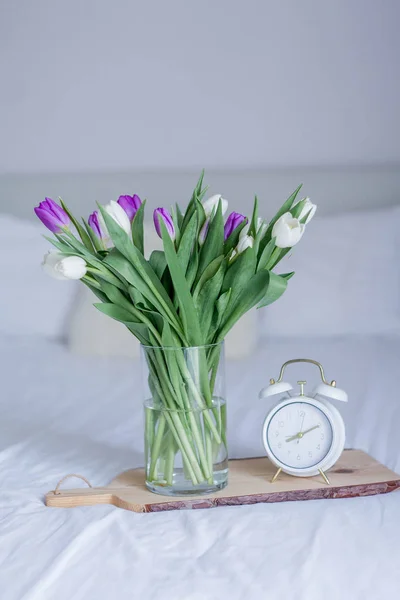 A bouquet of tulips in a glass vase, a white alarm clock on a wooden board stand on the bed. Spring. — ストック写真