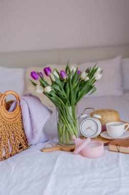 A bouquet of tulips in a glass vase, a white alarm clock, a candle, a note, a cappuccino mug on a wooden board are standing on the bed. Women's things. Breakfast in bed.