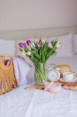 A bouquet of tulips in a glass vase, a white alarm clock, a candle, a note, a cappuccino mug on a wooden board are standing on the bed. Women's things. Breakfast in bed.