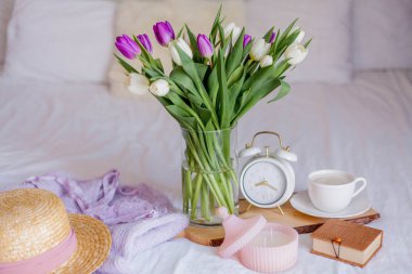 A bouquet of tulips in a glass vase, a white alarm clock, a candle, a note, a cappuccino mug on a wooden board are standing on the bed. Women's things. Breakfast in bed.