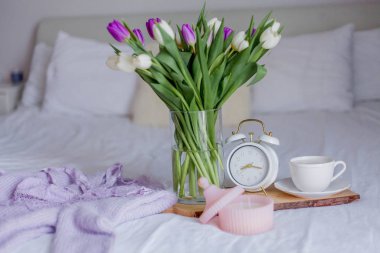 A bouquet of tulips in a glass vase, a white alarm clock, a candle, a note, a cappuccino mug on a wooden board are standing on the bed. Women's lace underwear. Breakfast in bed.