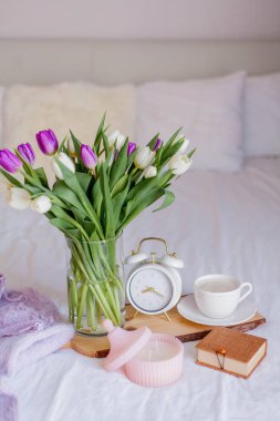 A bouquet of tulips in a glass vase, a white alarm clock, a candle, a note, a cappuccino mug on a wooden board are standing on the bed. Women's things. Breakfast in bed.