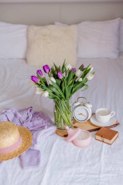A bouquet of tulips in a glass vase, a white alarm clock, a candle, a note, a cappuccino mug on a wooden board are standing on the bed. Women's things. Breakfast in bed.