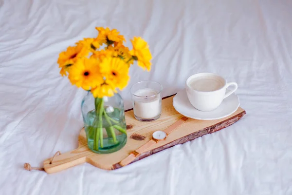 Een boeket gerbera 's in een glazen pot, een kopje cappuccino, een klok, een kaars op een houten statief liggen op het bed. Ontbijt op bed. — Stockfoto