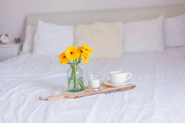Un ramo de gerberas en un frasco de vidrio, una taza de capuchino, un reloj, una vela en un soporte de madera están en la cama. Desayuno en la cama . — Foto de Stock