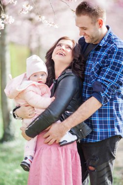 Genç ebeveynler kollarında küçük, güzel bir kız çocuğu tutuyorlar. Çiçekli bir bahçede güzel bir aile. Sakura. Bahar.