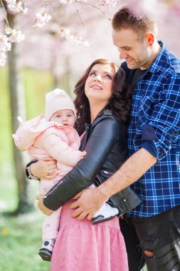 Genç ebeveynler kollarında küçük, güzel bir kız çocuğu tutuyorlar. Çiçekli bir bahçede güzel bir aile. Sakura. Bahar.