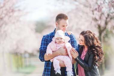 Genç ebeveynler kollarında küçük, güzel bir kız çocuğu tutuyorlar. Çiçekli bir bahçede güzel bir aile. Sakura. Bahar.